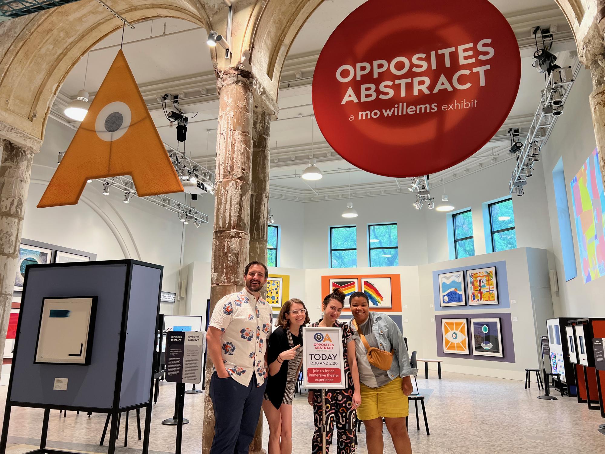 graduate students Michael DeWhatley, Claire Derriennic, Lily Odekirk and Renita James pose next to a sign, standing in front of the art exhibit OPPOSITES ABSTRACT: A MO WILLEMS EXHIBIT
