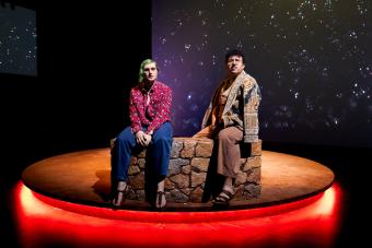 Two actors sitting on a stone bench look out into the distance, deep in thought, with stars projected behind them and a ring of orange light beneath them