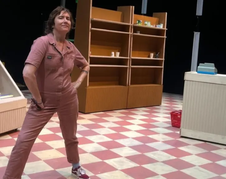 Photo of Lisa Damour standing in a pale red boiler suit on a checkered linoleum floor part of the grocery store set of her play frozen section