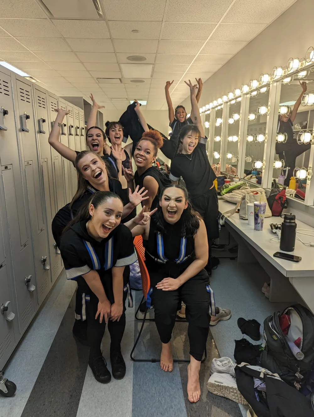 eight dancers pose with their arms raised excitedly in a dressing room with mirrors on one side and lockers on the other