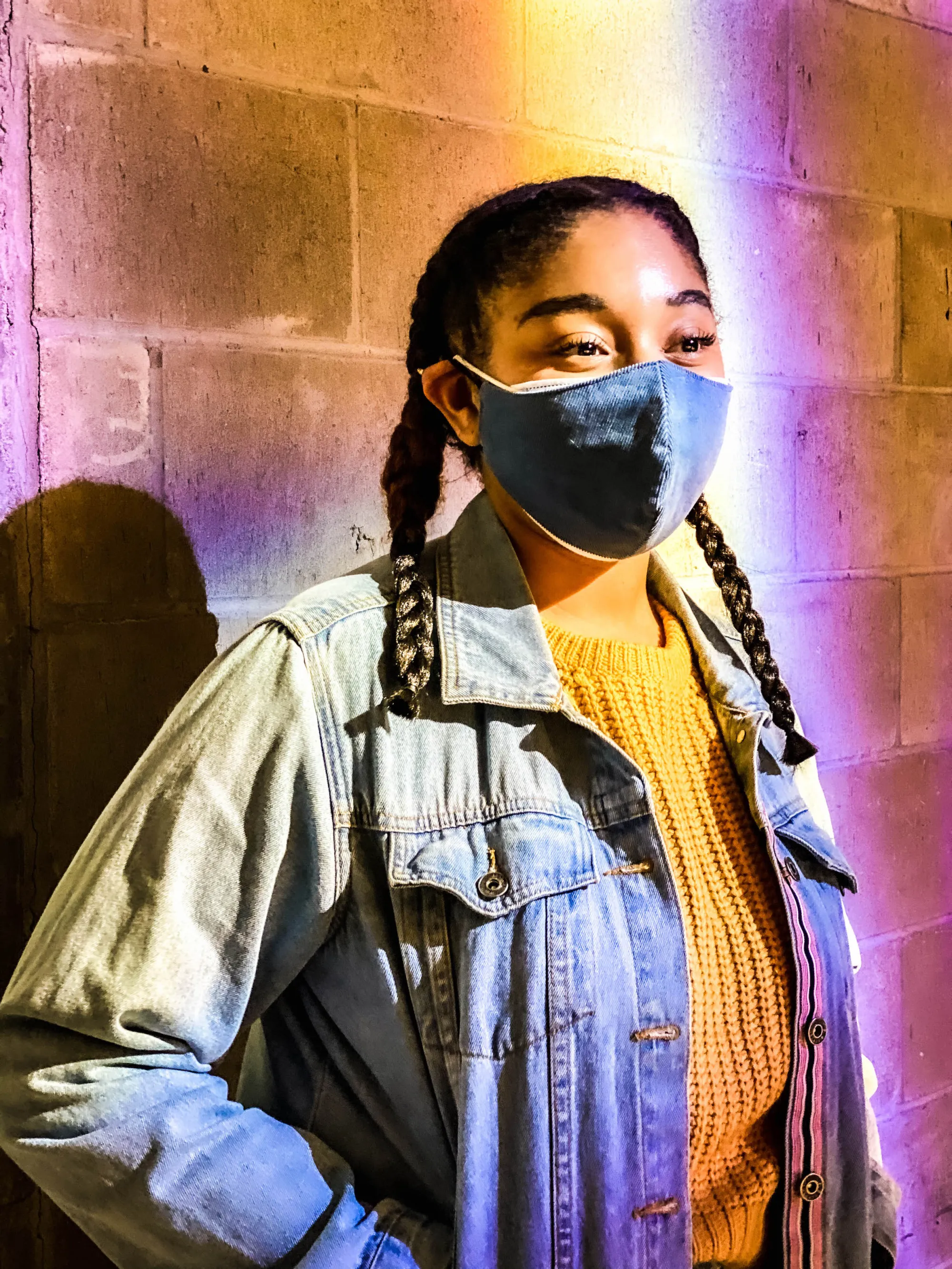 actress with her dark hair in braids wearing a mask, a yellow sweater and a blue jean jacket