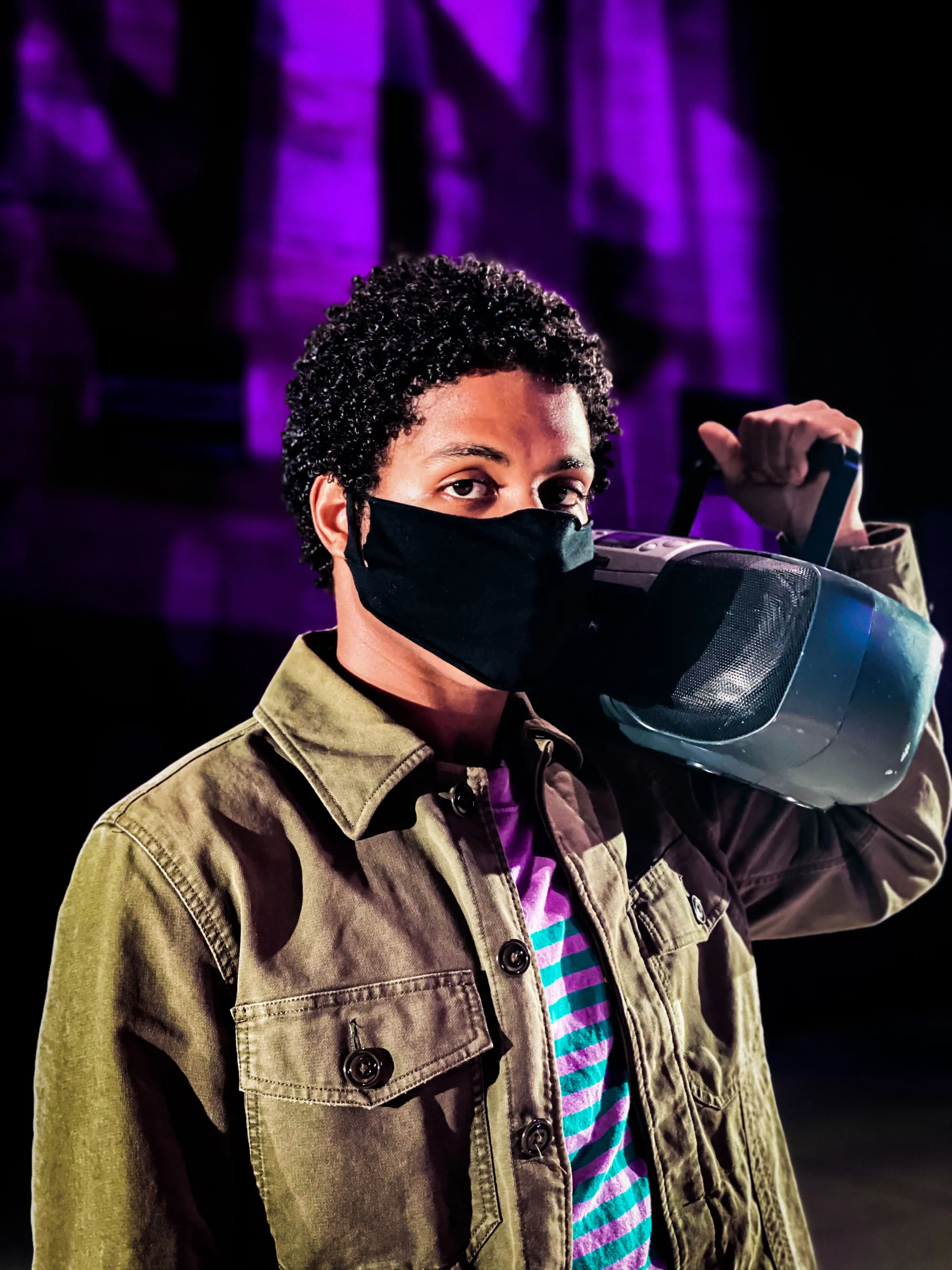 actor with short dark hair and a mask holds a radio on his shoulder