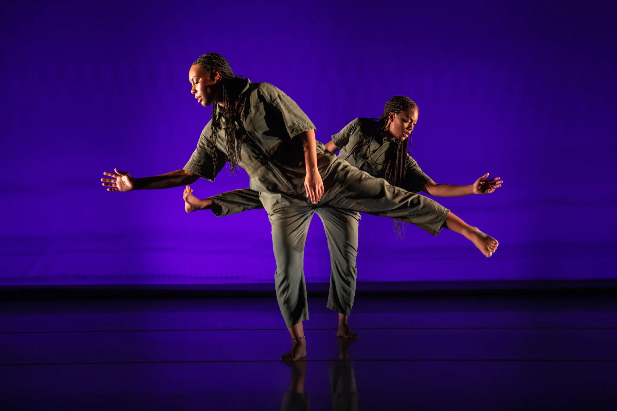 two dancers, one in front of the other, balance on one leg, holding their other legs up and reaching their arms out beside them