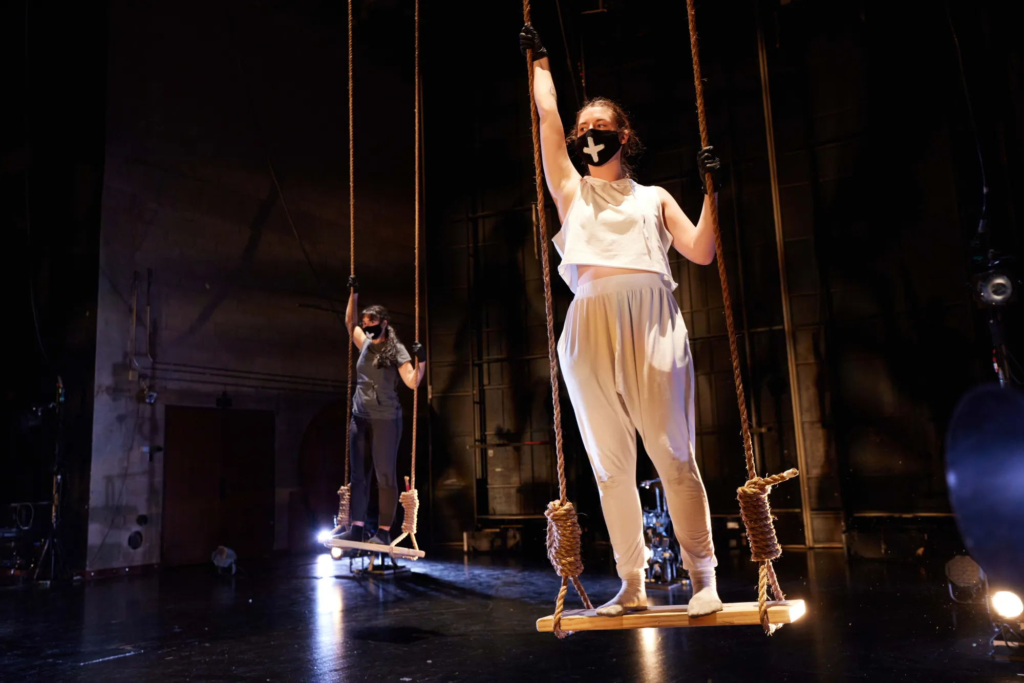 two dancers stand on wooden swings tied up by thick rope