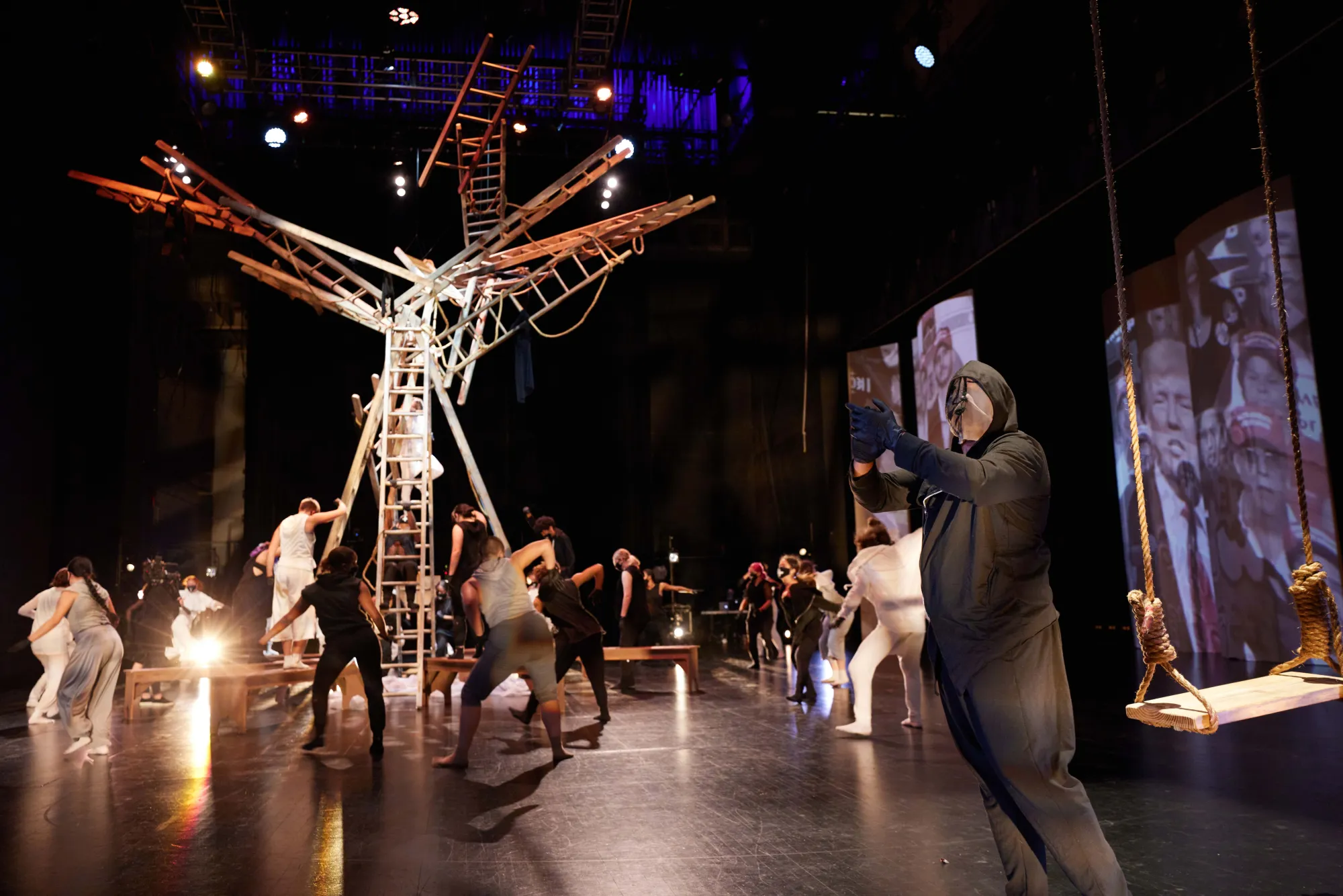 dancer in all black with a full face mask and a hood reaches his arm out, with dance ensemble moving behind him