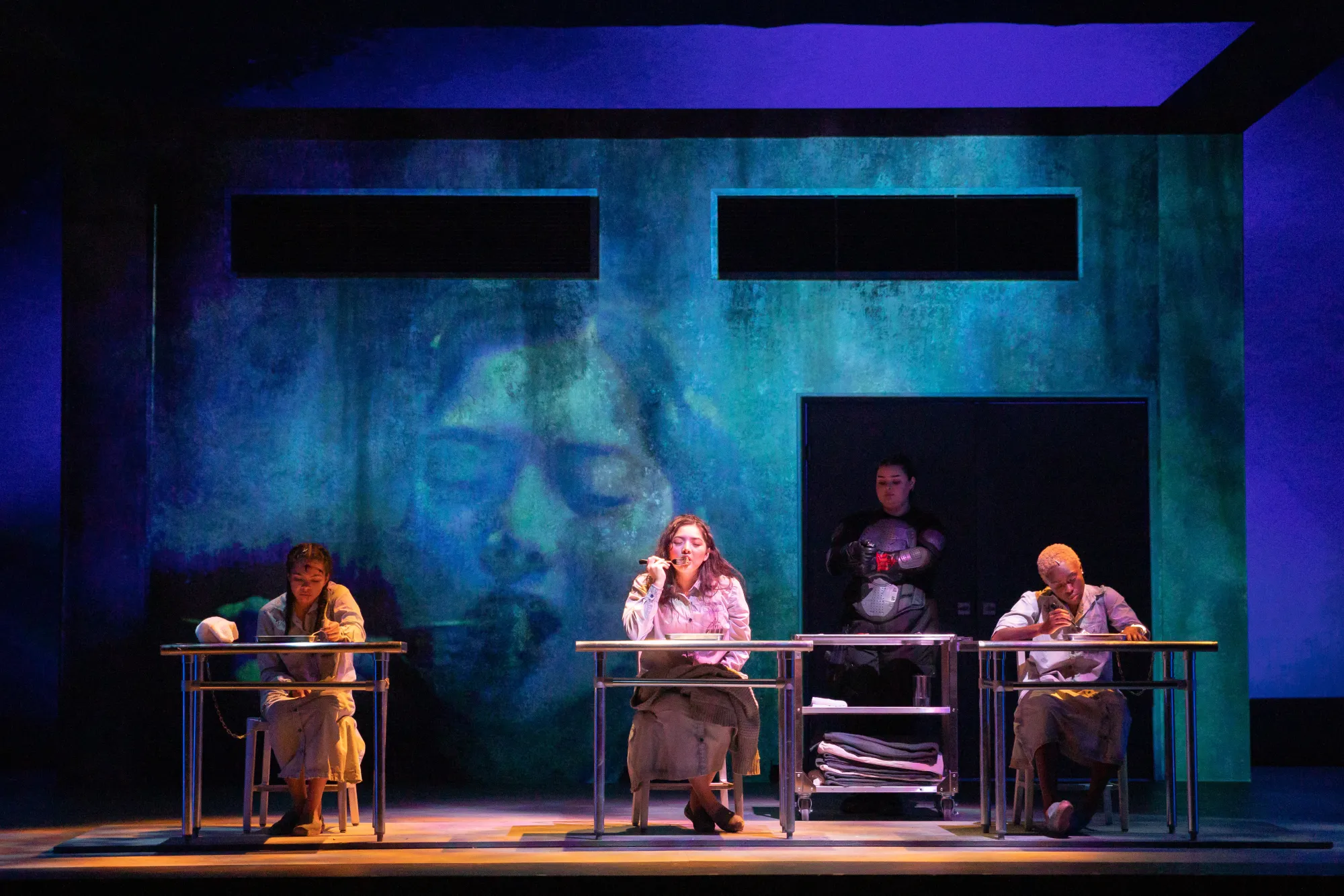 woman eats, with two other women sitting at tables next to her and a blue projection of her face in the background