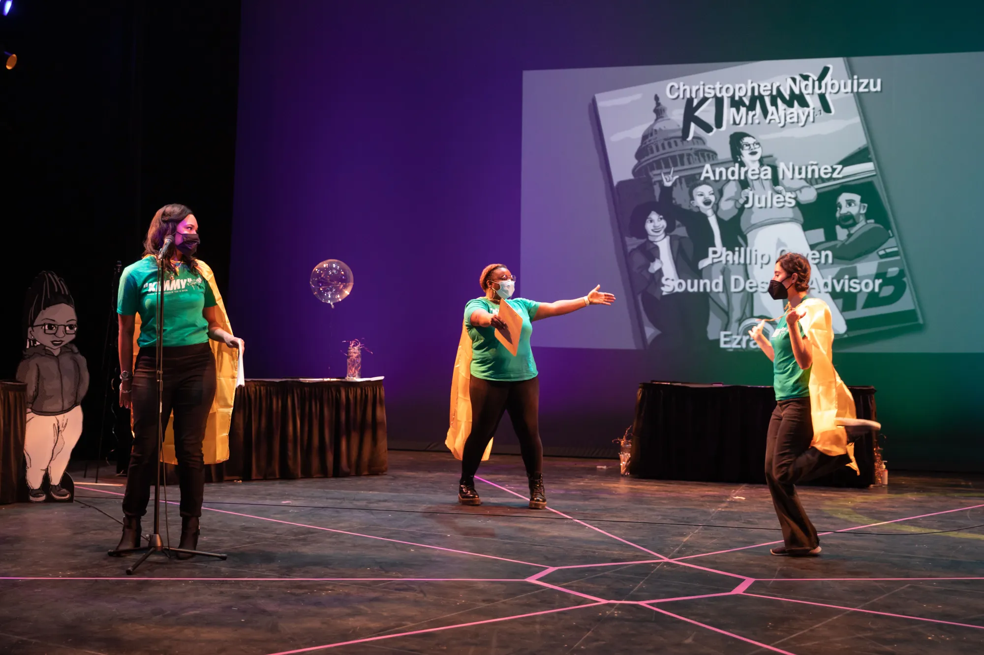 Two collaborators wearing yellow capes welcome a cast member, who is running onto the stage