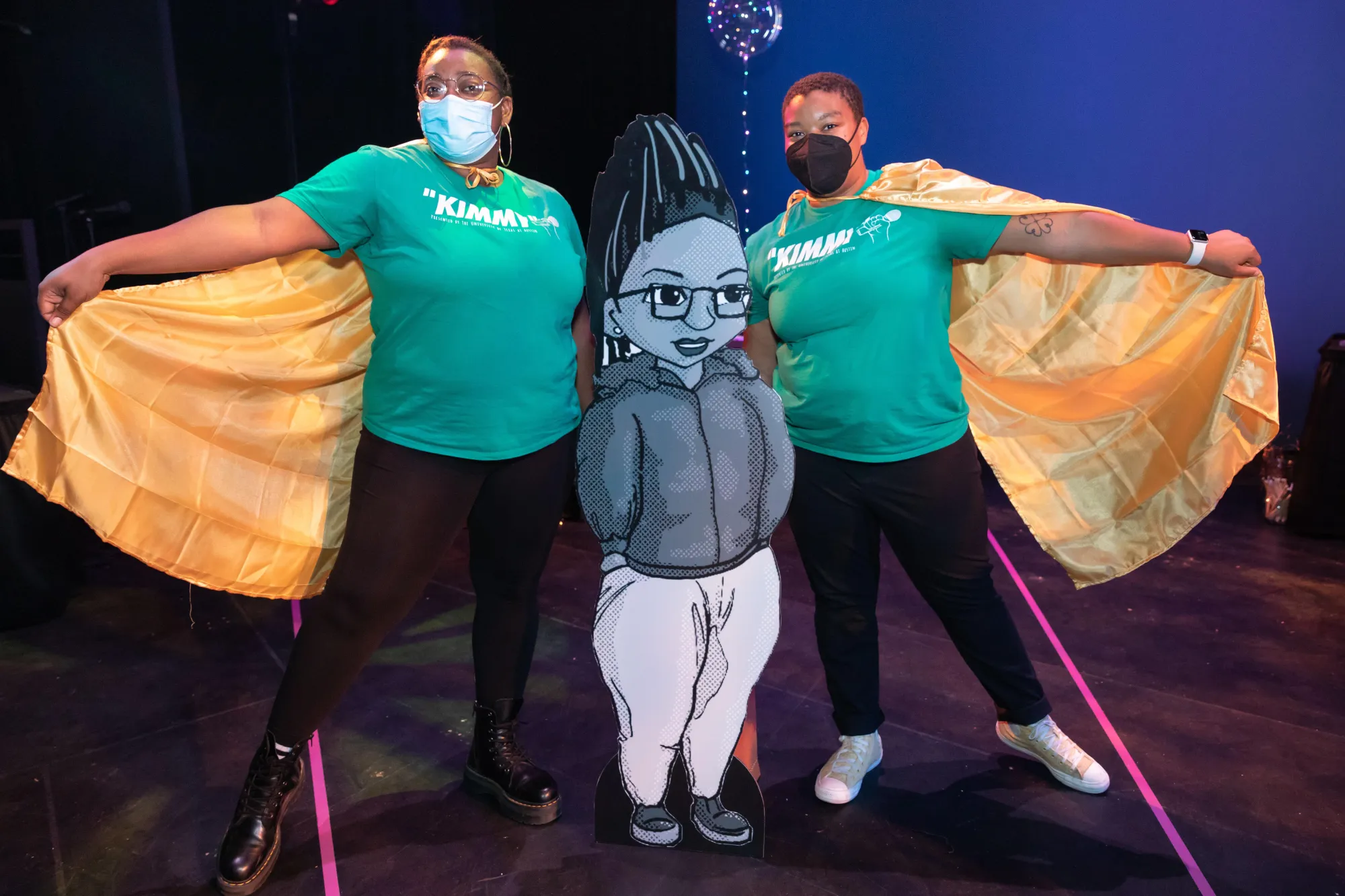 two collaborators hold up their yellow capes, standing on either side of a cardboard cutout of the character "Kemi"