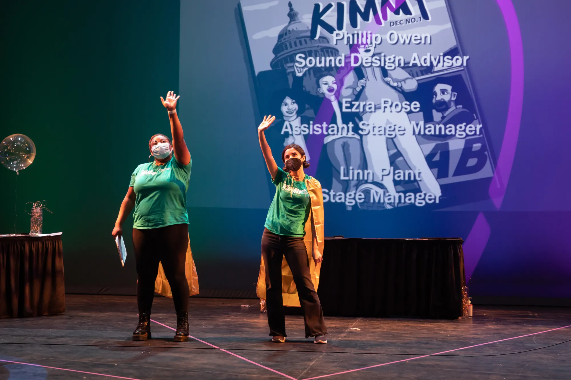 two cast members wearing green shirts and yellow capes wave at an audience, with a comic book projected behind them