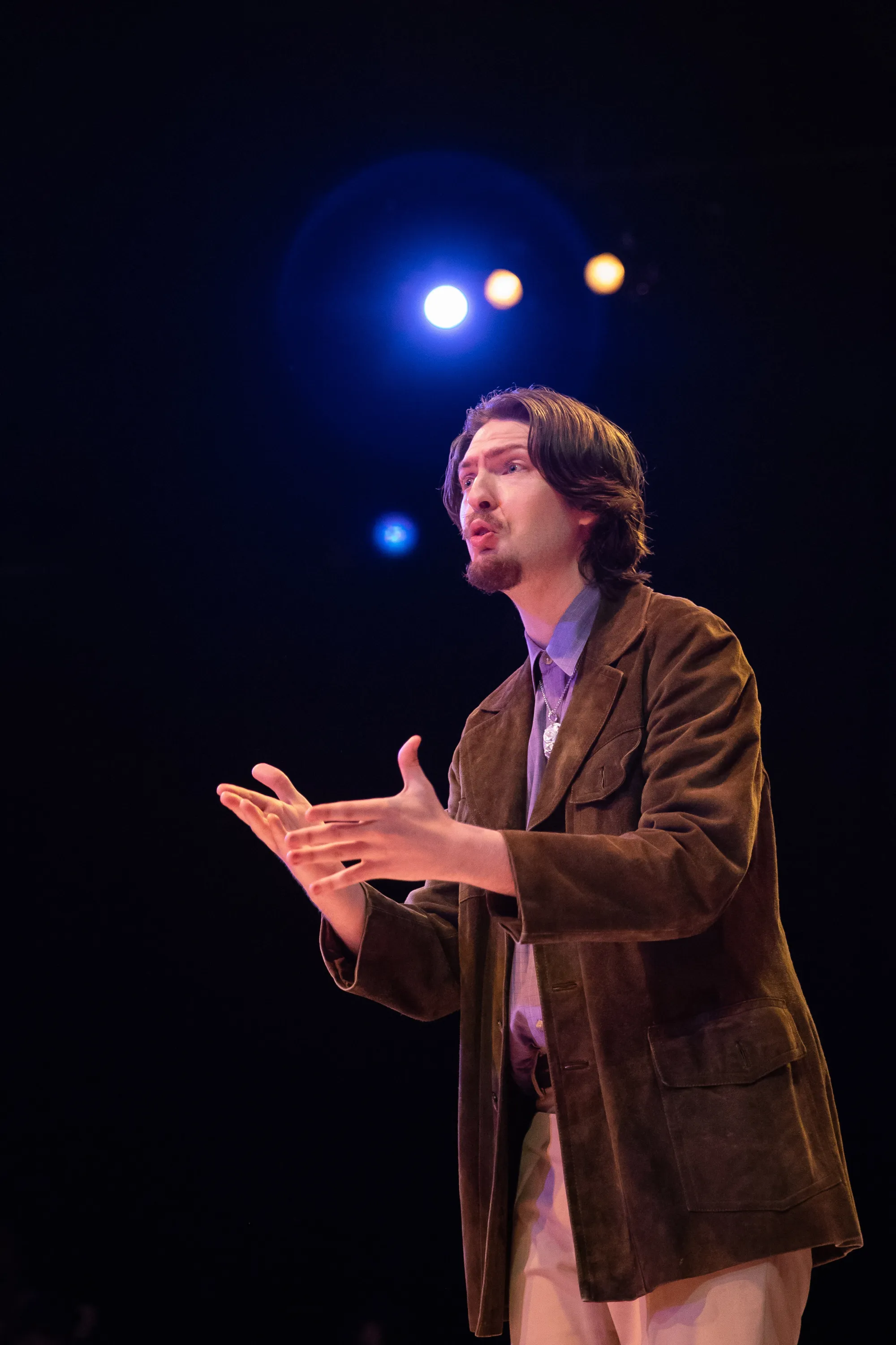 a man in a corduroy jacket speaks, holding his hands out, with stage lights shining behind him