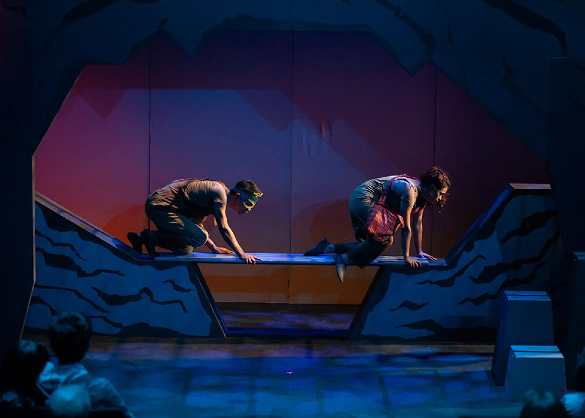 an actor in a mask and an actress carrying a bag crawl across a plank