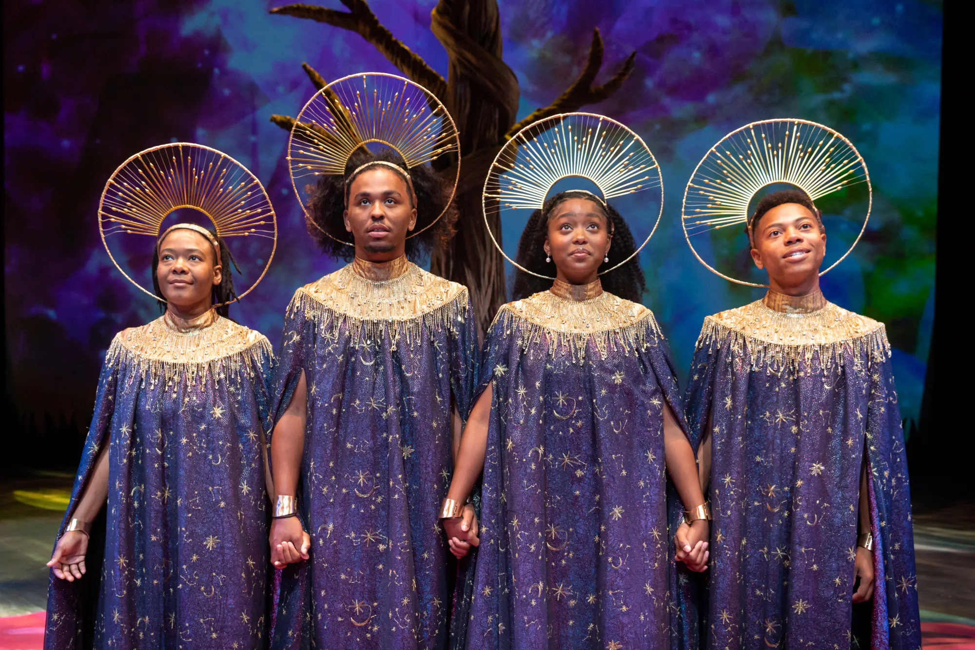 Four actors in purple and gold robes and gold halo-like head pieces hold hands, looking out hopefully into the distance