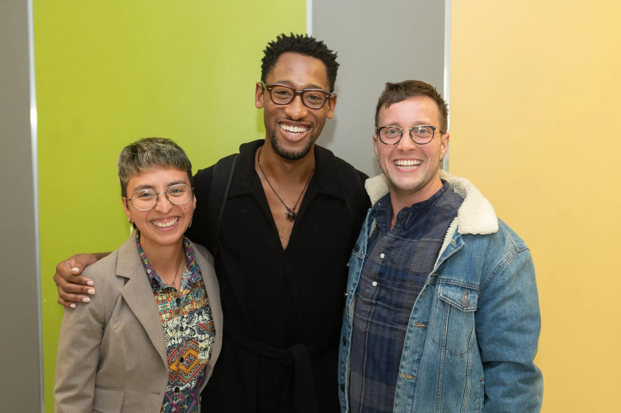 Three Performance as Public Practice students smile and pose together in front of a green, brown and yellow striped wall
