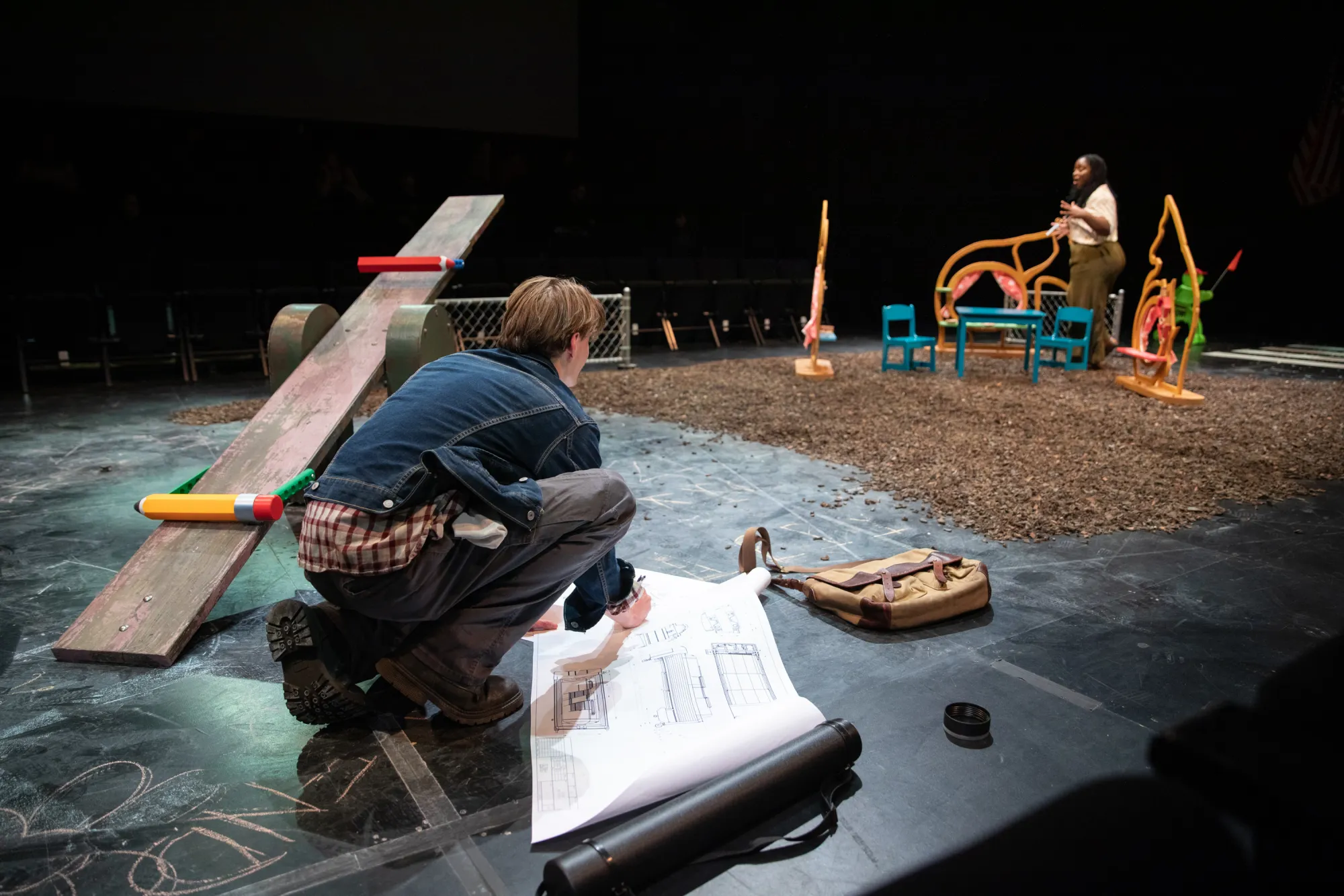 Actor Oliver Aaro as Arthur crouches down in the corner of the stage as he unfurls his plans for new playground equipment. Taken from his perspective on the floor, this view shows the on-stage playground and actress Shalom Onuorah as Jen in the background.