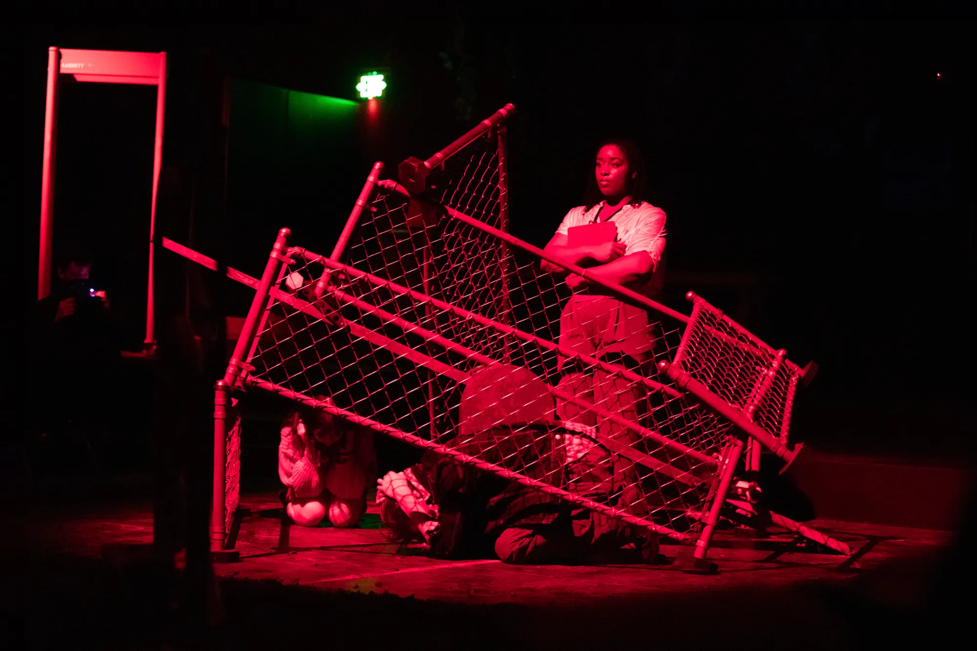 Shalom Onuorah as Jen stands center stage basked in red light as Penny Lou Zimmerman as Clara and Oliver Aaro as Arthur are curled up on the ground hiding. They are performing a gun violence drill.