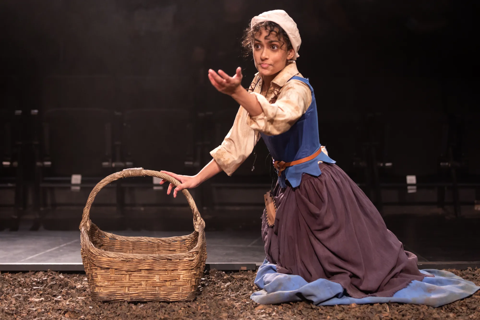 Carlise Rosa as Frau Thera kneels on the potato field with her right hand on the handle of her basket and her other gesturing towards the audience.