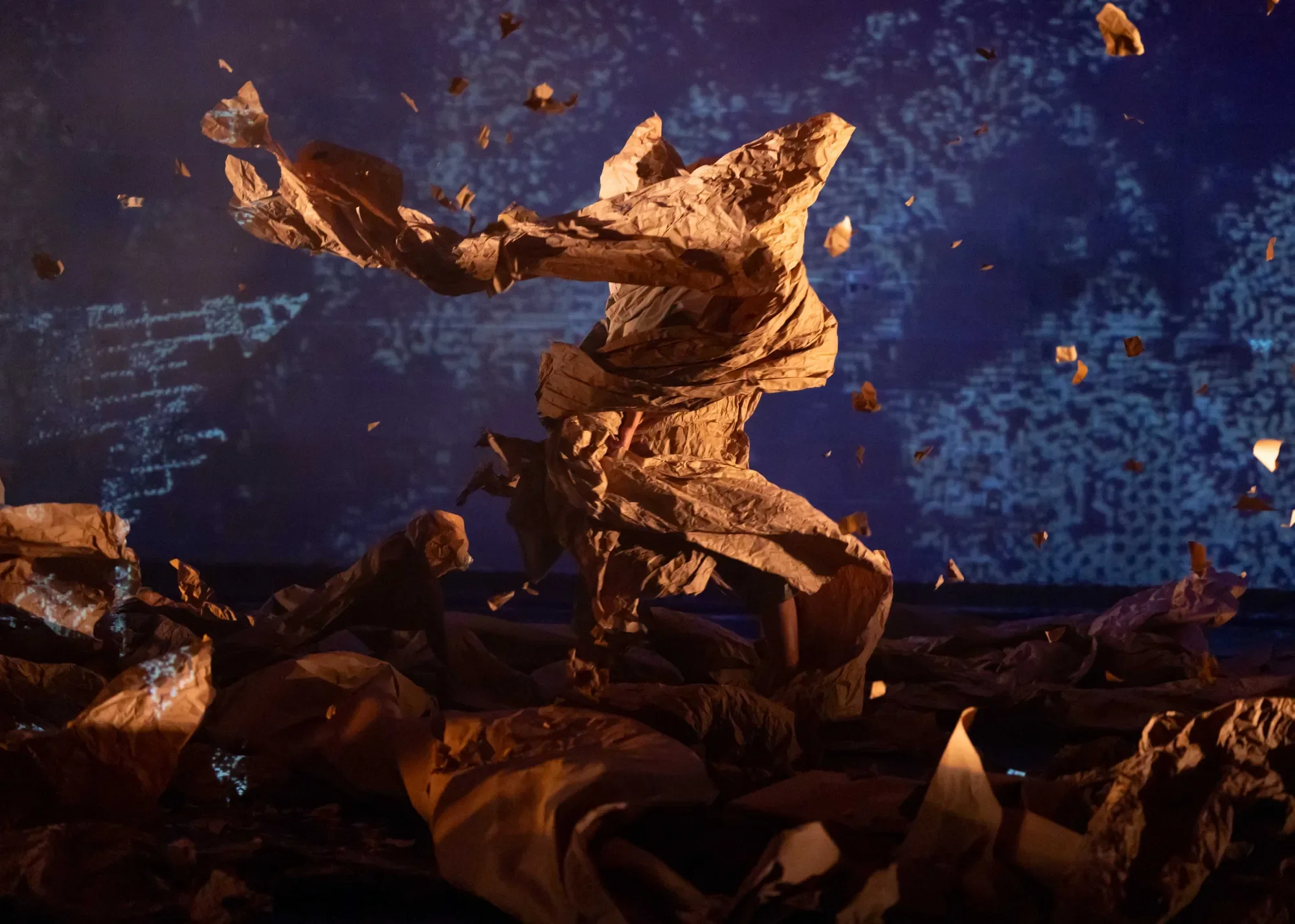 Long brown paper that once was straight is crumbled on the stage, with dancers bodies interspersed. A large sheet of crumpled paper spirals upward in the wind tunnel of recent movement.