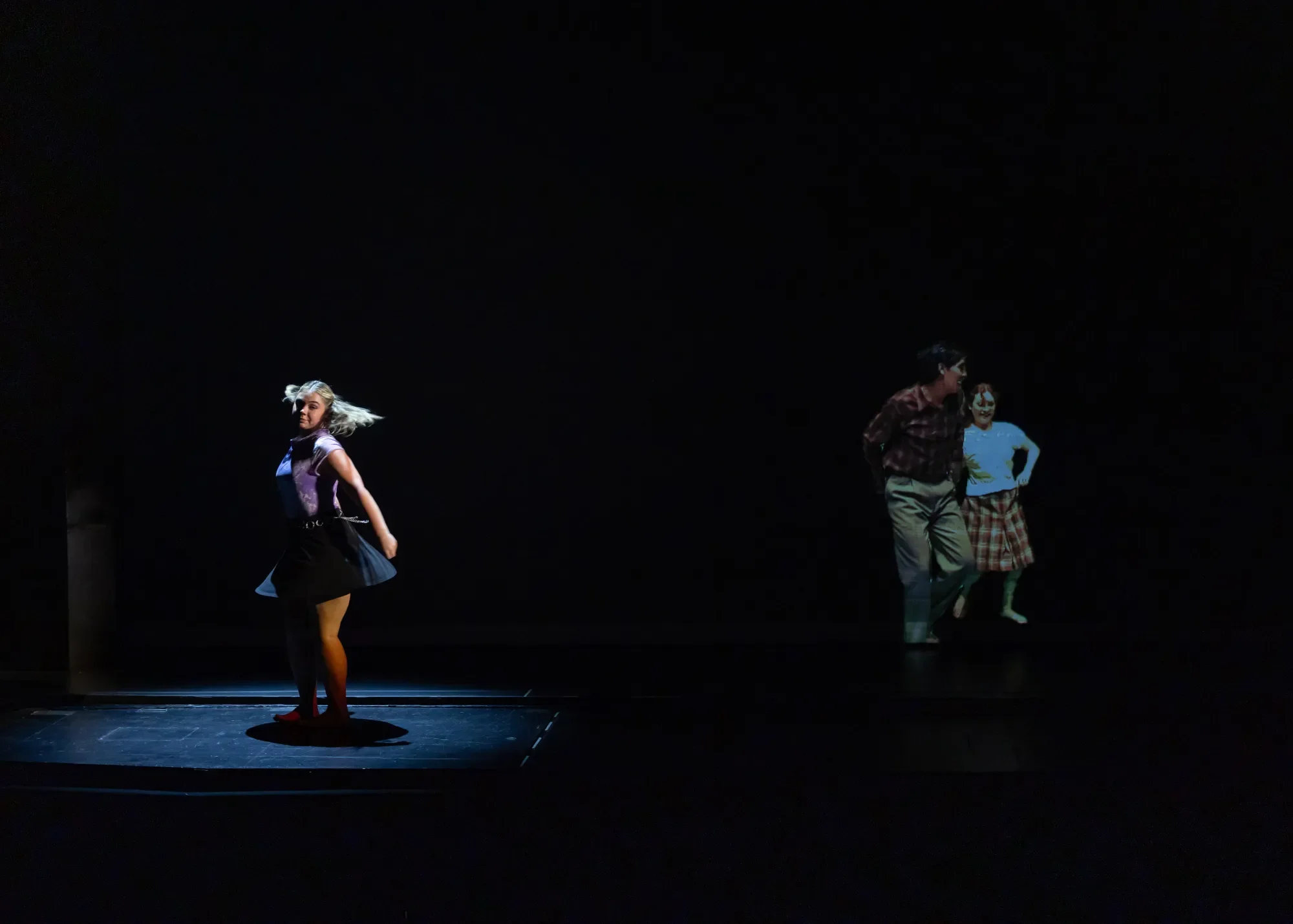 A dancer twists in a spotlight on stage, with two other dancers projected on a scrim next to her