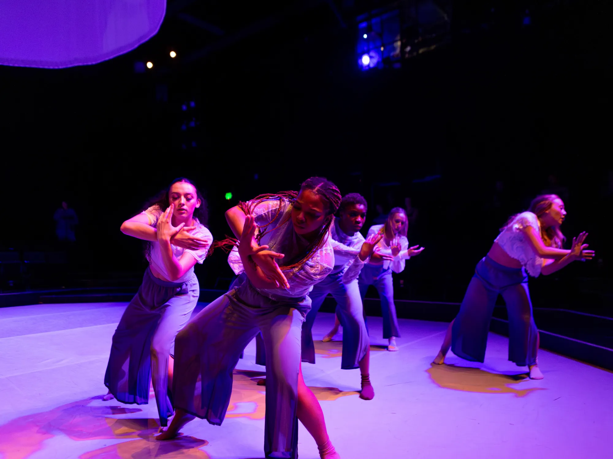 Dancers are basked in purple light, emotion ripe on their faces as they cross one wrist over the other and bend their knees.