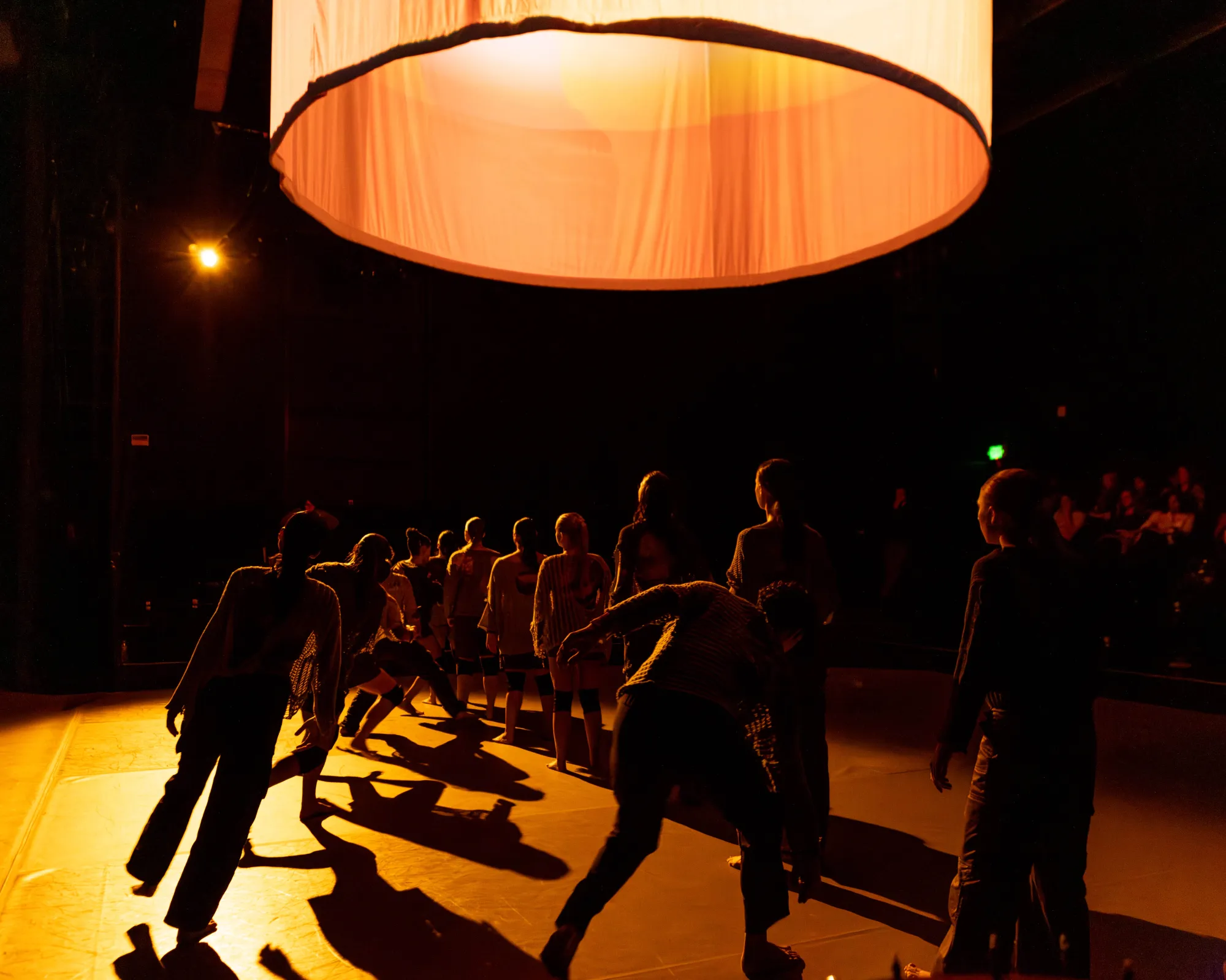 Cloaked in darkness, standing against a dimmed light, dancers are lined up as some move in and out of the diagonal they are standing in.