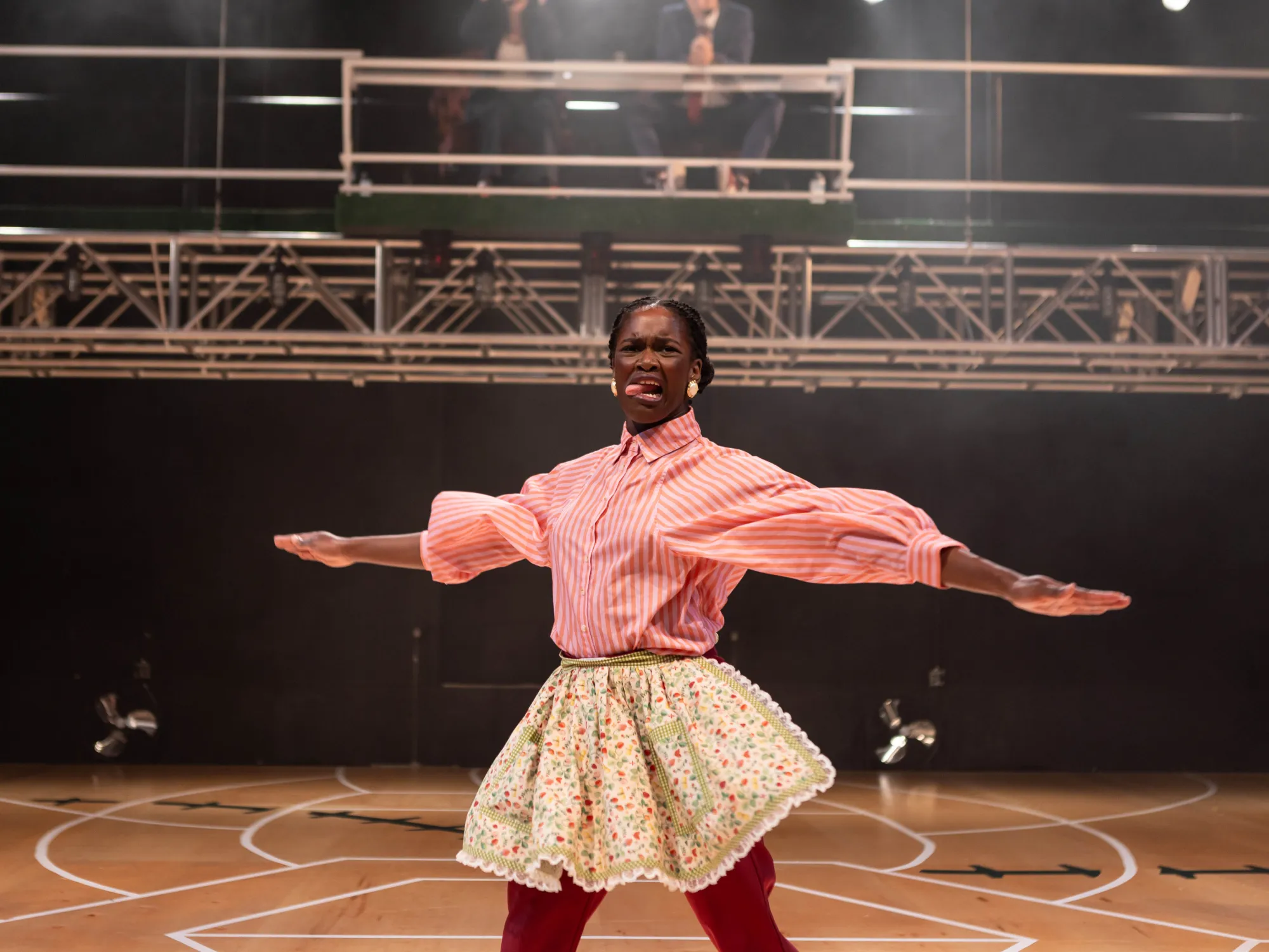 The oldest sister, Trifle, played by Oyin Ojomo swings her arms from side to side, sticking her tongue out. She is depicting the sport of cleaning up dishes.