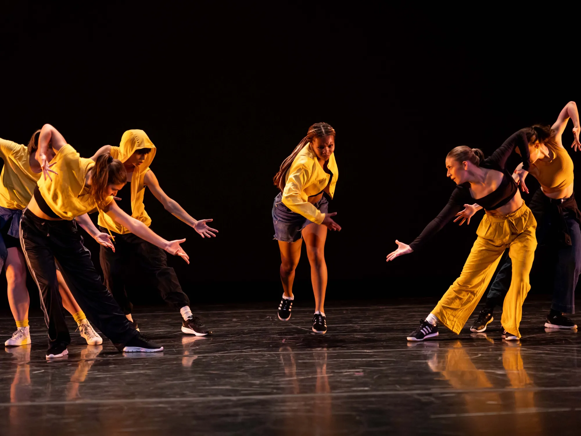Dancers cloaked in black and yellow in Lone. Lean. Less. lean towards the center of the stage with their arms in a diagonal.