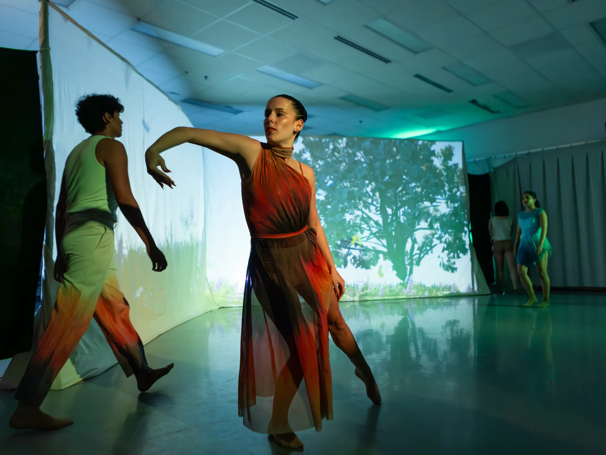 An undergraduate dancer leans on their right leg and reaches forward, with a projection of trees behind her
