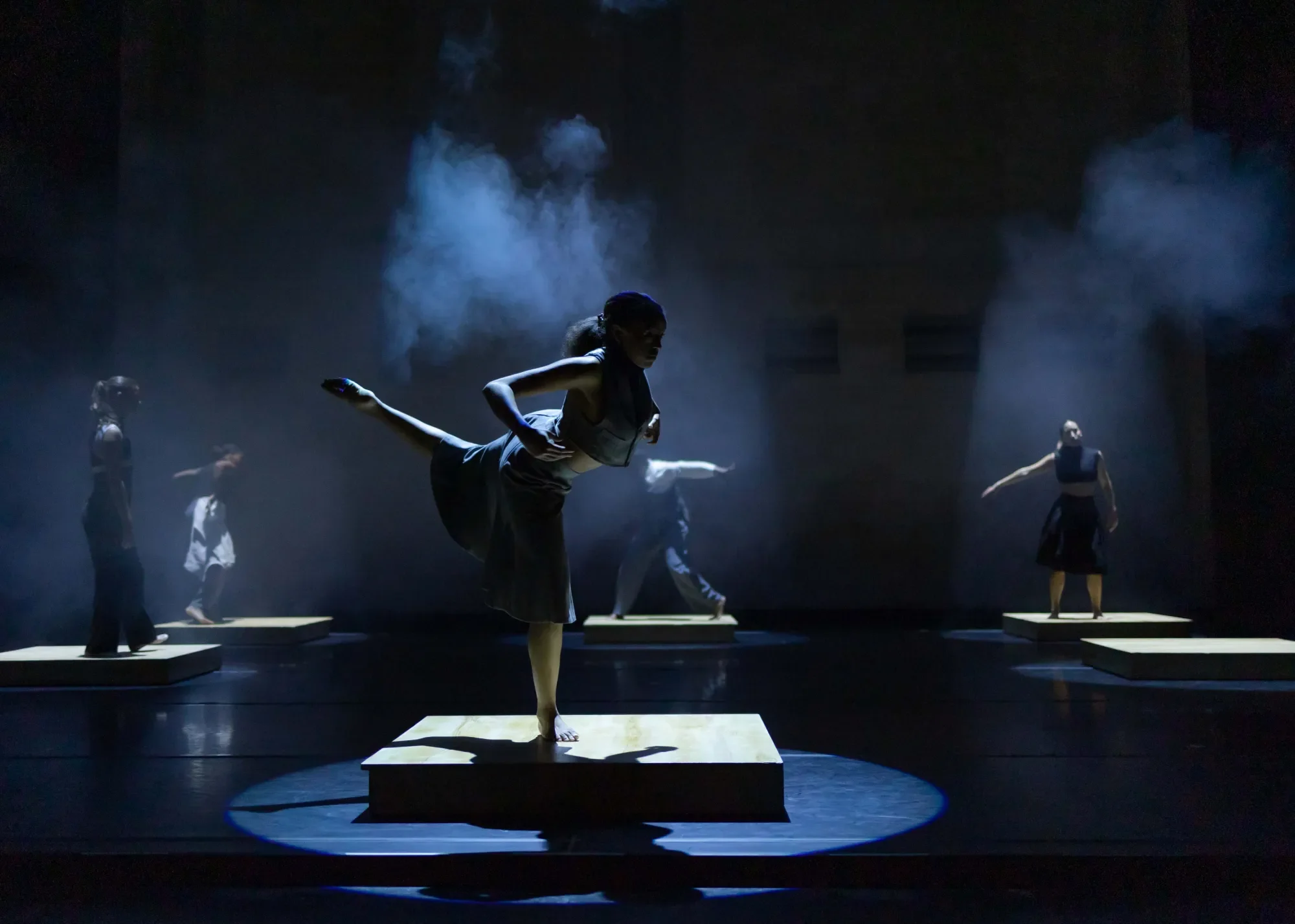 A dancer poses on a platform in a spotlight, standing on their right foot and pointing their left leg behind them