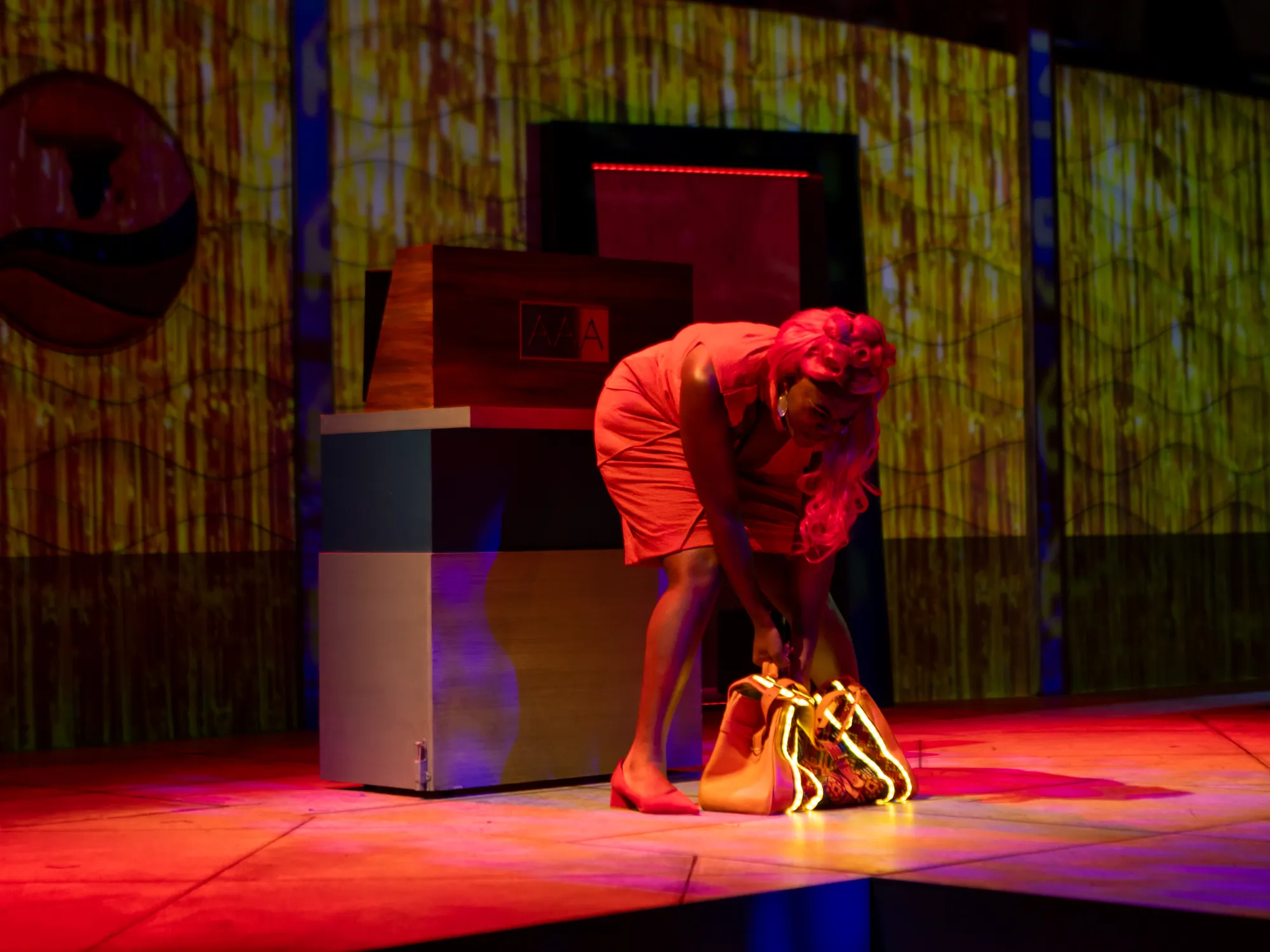 An actor in a red dress reaches to lift up a bag