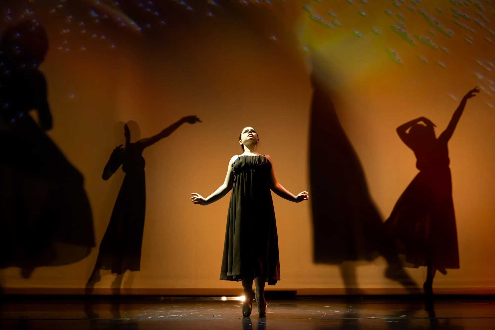 A dancer stands en pointe in front of three shadows
