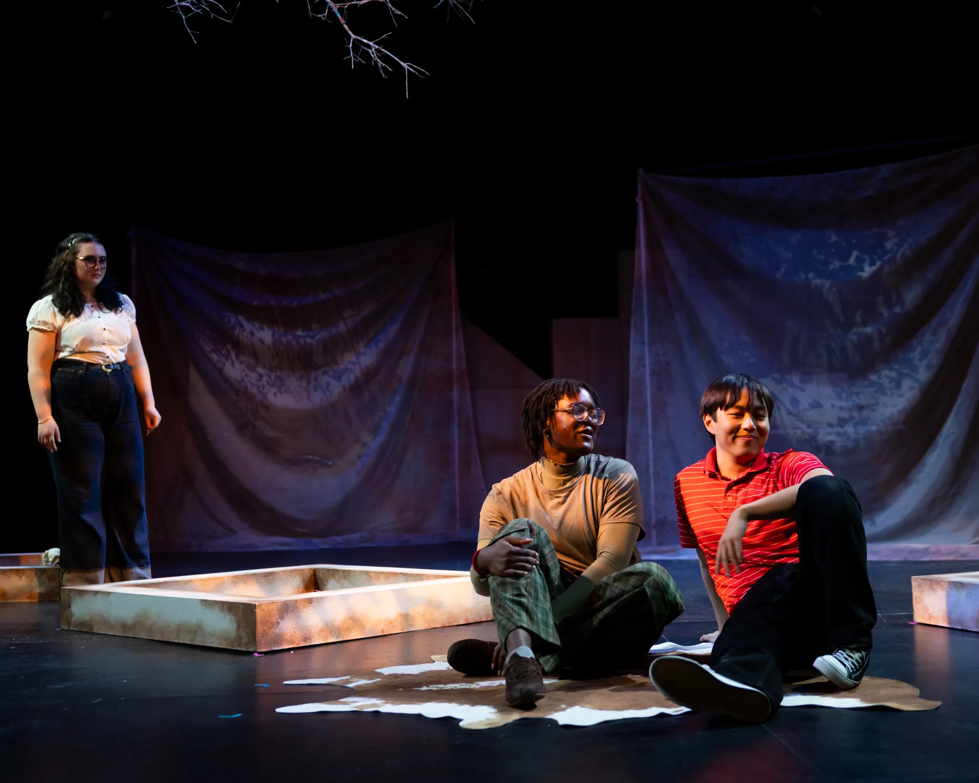 Two actors sit on a cow skin rug