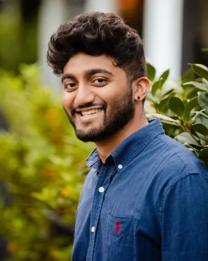 A man in a dark blue collared shirt standing in front of leaves
