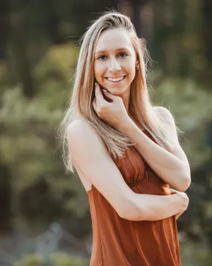 A woman with long blonde hair touches her cheek and smiles