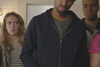 man with a beanie and a zip-up hoodie talks, with a woman and a man watching him in the background