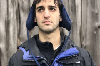 man with short brown hair wearing a blue hat and jacket and standing in front of a wooden fence