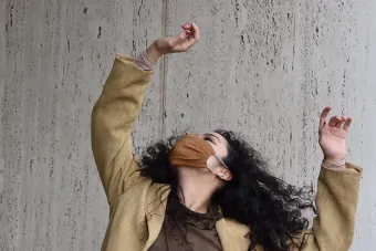 dancer with a tan coat and mask on posing with her arms up in front of a white stone wall