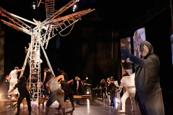 dancer in all black with a mesh mask over his face reaches forward, with other dancers in the background around a set piece that looks like a tree made out of ladders