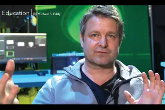 a man with blue eyes and graying hair speaks using his hands, with a computer screen and other integrated media elements behind him