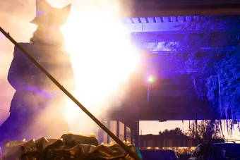 actor performing as Hades on an outdoor stage, spotlighted in bright, warm light with cars and blue lighting in the background