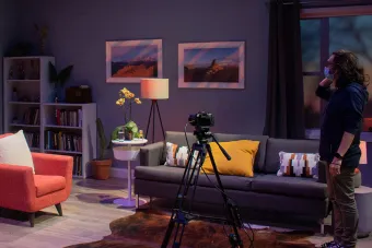 man with long brown hair and a mask stands on a set with a couch, chair, a cowhide rug and a camera