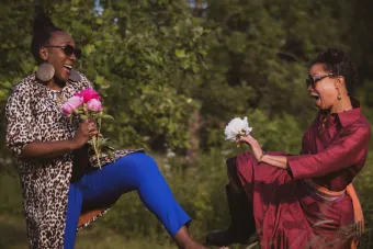 two women wearing sunglasses and holding flowers pose with one of their legs lifted