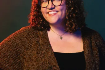 woman with brown curly hear smiles, wearing glasses and a brown knitted cardigan