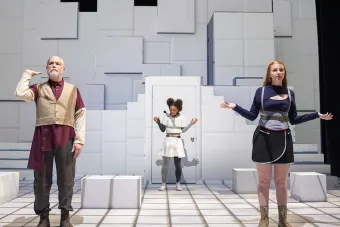 three actors stand on stage, one holding up his arm as if holding an imaginary tray, the other two holding their arms up and shrugging