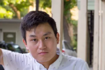 man with short black hair and a white, patterned button up leans on a column with his right arm
