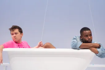two men sit in a bathtub wearing a pink and blue shirt and holding two balloons