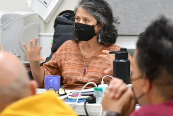 woman wearing blue earrings and a black mask gestures to a room of actors
