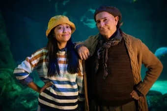 A woman wearing a striped nautical t shirt and a fisherman's cap smiles alongside a gruff but kind mariner