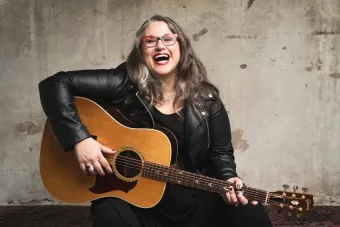 Headshot for alumna and Producing Artistic Director of Oregon Children's Theatre Jenn Hartmann Luck, who's smiling and holding her guitar