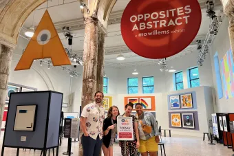 graduate students Michael DeWhatley, Claire Derriennic, Lily Odekirk and Renita James pose next to a sign, standing in front of the art exhibit OPPOSITES ABSTRACT: A MO WILLEMS EXHIBIT