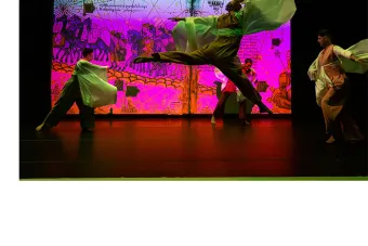 Dancer mid-jump, their sleeves flowing out beside them, with other dancers and an intricate projection of historical art and maps in the background